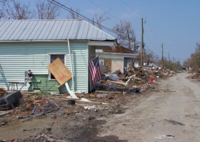A residential street shows homes surrounded by storm debris, with broken boards, scattered household items, and mud covering the ground. An American flag hangs from one damaged house amid the destruction.