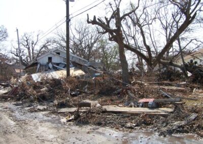 A muddy residential street is filled with wreckage, including collapsed fences, broken lumber, and uprooted trees. Houses in the background show significant structural damage.