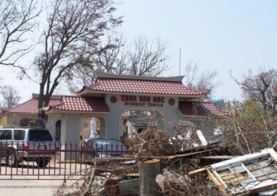 A Buddhist temple with a red roof stands amid heavy storm debris piled along the street, including tree limbs, broken boards, and window frames. Damaged trees surround the building.