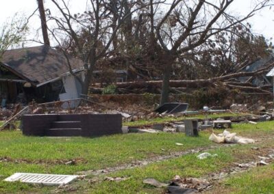A neighborhood is filled with downed trees, broken structures, and scattered debris, with only concrete steps remaining from a destroyed home in the foreground. The surrounding trees are stripped bare from the storm.