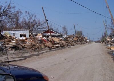 A dirt road runs between homes heavily damaged by Katrina, with collapsed walls, torn roofs, and scattered debris filling the yards. Power lines lean overhead along the devastated street.