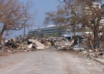 A road is blocked by massive piles of storm debris, including shattered wood, roofing, and metal, with a damaged multi‑story building visible in the background. Bare trees line the sides of the roadway.