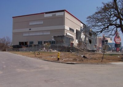 A damaged commercial building with torn exterior walls and missing sections sits near a fire hydrant and empty street. Nearby structures and trees show widespread storm destruction.