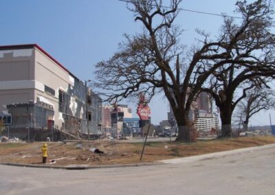 A large commercial building stands with extensive façade damage, missing panels, and broken walls, surrounded by a barren landscape of dead trees. Nearby signs and fencing lean at odd angles from the storm.
