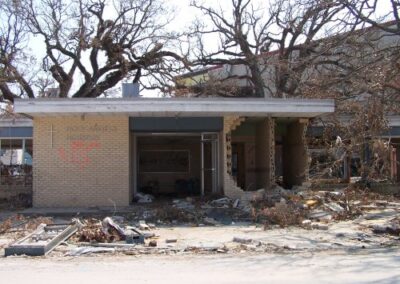 A brick building with its front walls blown out sits amid scattered rubble, broken furniture, and tangled branches. Spray‑painted markings and exposed interior rooms show the extent of hurricane damage.