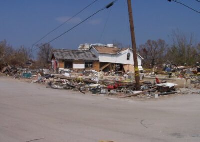 A storm‑ravaged home sits amid a massive field of debris, with roofing, lumber, and household items scattered across the property. A leaning utility pole stands beside the damaged structure under a clear sky.