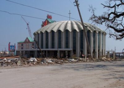 A large domed church building stands with visible exterior damage and debris piled around the base, while a crane operates in the background. Leaning utility poles and stripped trees surround the scene.