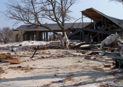 A collapsed residential structure lies in pieces across a concrete slab, with bricks, broken beams, and roofing spread throughout the yard. A large tree trunk lies uprooted among the destruction.
