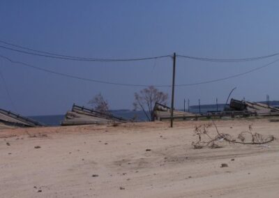 Large concrete bridge sections lie collapsed along the shoreline, with leaning utility poles and scattered debris on the sandy ground. The calm water contrasts with the severe structural destruction.