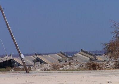 Massive concrete bridge spans lie toppled in sections along the shoreline, with dirt, rubble, and a leaning power pole in the foreground. The water is visible just beyond the collapsed structure.
