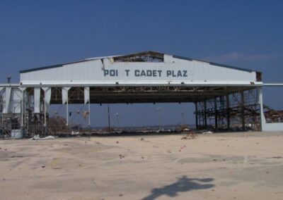 A heavily damaged waterfront pavilion labeled “POINT CADET PLAZ” stands mostly open on all sides after losing walls and roof panels. The surrounding pavement is littered with broken metal and debris.
