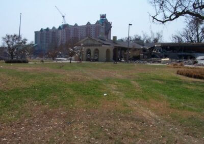 A grassy park area stretches toward a damaged pavilion, with a tall hotel building visible in the background. Wreckage and bare trees line the edges of the park.