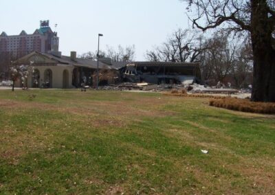 A grassy park area faces a destroyed pavilion and surrounding rubble, with bare trees and a tall hotel visible in the background. Storm debris remains scattered near the building edges.