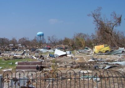 A wide field of post-storm destruction is filled with mangled debris, broken structures, and uprooted trees, with a blue water tower rising in the distance. Shredded metal and scattered materials cover the ground.