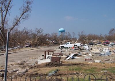 A devastated neighborhood is littered with splintered lumber, broken appliances, and overturned vehicles, with a blue water tower rising in the background. Bare, damaged trees line the scene.