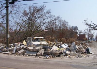 A crushed vehicle sits amid a massive pile of concrete slabs, twisted metal, and broken debris along a storm‑damaged roadside. Bare trees and scattered wreckage fill the background.