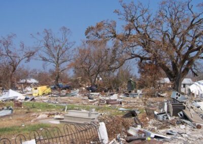 A wide field of destruction is filled with shattered structures, broken fencing, and debris scattered among bare trees. A partially collapsed yellow vehicle sits in the center of the wreckage.