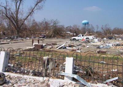Storm‑ravaged lots stretch across a neighborhood with scattered debris, damaged vehicles, and uprooted trees under a blue sky. A fence in the foreground is bent and tangled with broken materials.