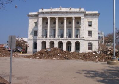 A historic white building labeled “CITY HALL” stands behind a large mound of storm debris piled along the front walkway. The structure’s tall columns remain intact despite the surrounding destruction.