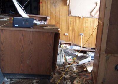 A demolished library interior with piles of broken drywall, insulation, and debris scattered across the floor. Portions of the remaining walls show exposed studs and storm damage.