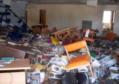 A library room is filled with collapsed furniture, mud‑soaked books, broken shelves, and debris scattered across the floor. Several chairs and tables are overturned amid the destruction.