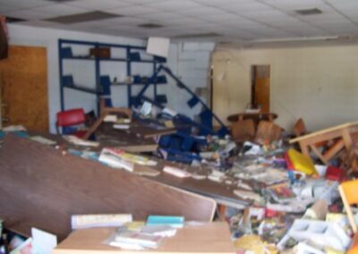 A heavily damaged library interior shows books, shelving, and furniture pushed into chaotic piles across a mud‑covered floor. Walls and ceiling panels are warped or missing, exposing structural areas.