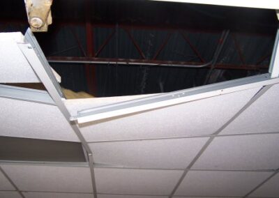 A close‑up view of a ceiling shows a broken grid with tiles missing, revealing metal roofing beams and insulation above. The damaged ceiling panels hang loose from their frame.