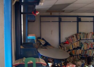 A children’s library area with blue wall shelves shows piles of warped and water‑damaged books on the floor and a round table pushed to the side. The ceiling tiles are sagging or missing due to storm damage.