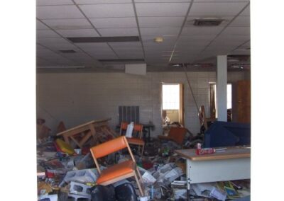 A library room is filled with mud‑covered debris, overturned furniture, scattered books, and a damaged ceiling with broken tiles. Orange chairs and tables lie toppled among ruined materials across the floor.