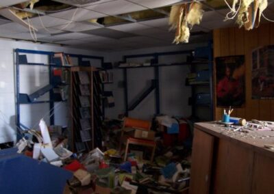 A damaged children’s reading area shows toppled shelves, scattered books, broken furniture, and ceiling debris littering the floor. The wall shelving is bent and partially detached from storm force.