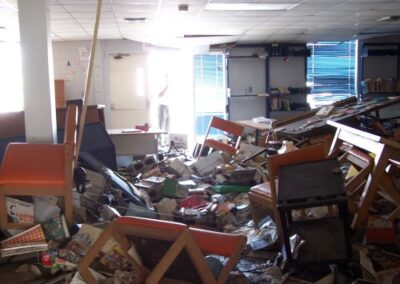 A library room filled with overturned chairs, desks, and debris is lit by bright sunlight coming in through an open doorway. Books, papers, and storm‑damaged materials are piled across the mud‑covered floor.