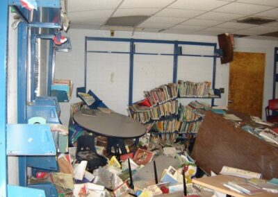 A close‑up of the floor shows a large collection of soaked children’s books, debris, and damaged storage containers scattered throughout the area. Mud and water stains cover nearly every surface.