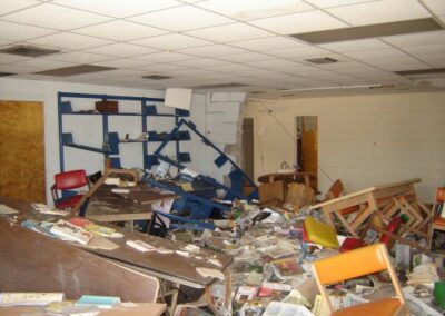 A children’s library area shows blue shelving units on the wall with books collapsed into piles below, mixed with debris and water‑damaged materials. Ceiling tiles above are broken or missing from storm impact.