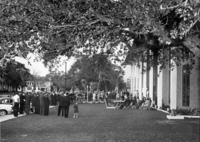 A wide shot of the dedication ceremony showing attendees seated on the lawn, a speaker at the podium, and groups of spectators gathered near the sidewalk beneath large overhanging tree branches.