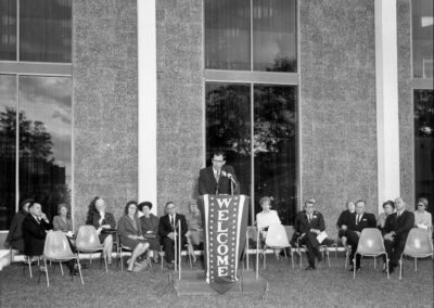 A speaker stands at a podium with a “WELCOME” banner, surrounded by seated guests arranged in rows in front of the library’s tall windows.