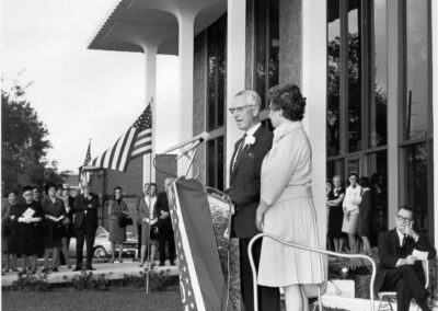 A dedication ceremony taking place outside the library, with two speakers standing at a podium draped in patriotic fabric while a crowd and color guard stand nearby. The building’s tall modern columns and large windows form the backdrop.