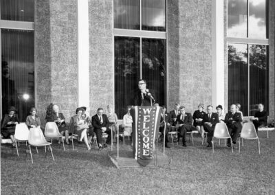 A speaker addresses the audience from a podium marked “WELCOME,” surrounded by formally dressed guests seated in front of the library’s tall windows.