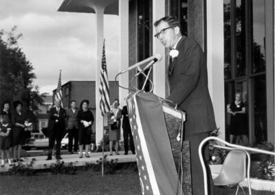 A speaker stands at a podium in front of the library’s modern glass-and-column exterior, with American flags displayed behind and attendees standing in the background.
