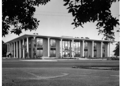 A black‑and‑white photograph of a modern two‑story library building featuring tall vertical columns, large floor‑to‑ceiling windows, and landscaped grounds, viewed from across the street with tree branches framing the scene.