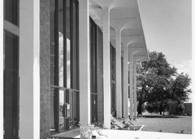 A black‑and‑white photograph of a modern library building’s exterior featuring tall vertical columns, floor‑to‑ceiling windows, landscaped greenery, and a narrow reflecting pool beside a walkway.