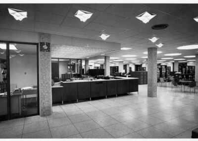 A black‑and‑white photograph of a spacious library interior featuring a long central service desk, tiled floors, textured support columns, overhead geometric light fixtures, bookshelves, and tables with chairs.