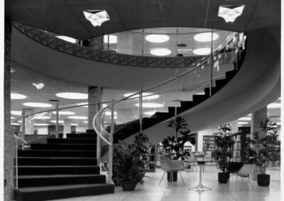 A black‑and‑white photograph of a sweeping spiral staircase inside a library, with carpeted steps, glass railings, potted plants, and seating arranged beneath the upper level’s circular balcony.