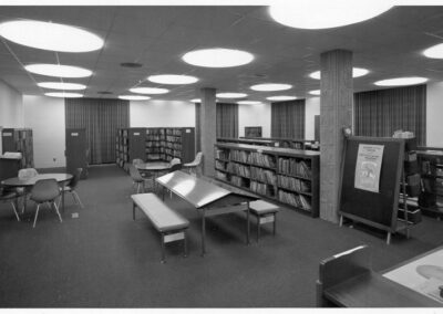 A black‑and‑white photograph of a library interior showing low bookshelves filled with materials, round tables and chairs, long benches around a slanted display table, and textured support columns beneath large circular ceiling lights.