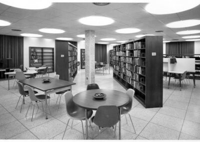 A black‑and‑white photograph of a library interior featuring rows of tall bookshelves, round and rectangular tables with molded plastic chairs, tiled floors, and large circular ceiling lights.