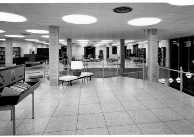 A black‑and‑white photograph of a library’s upper floor showing tiled floors, textured support columns, glass railings overlooking the lower level, bookshelves, and seating areas arranged beneath large circular ceiling lights.