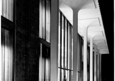 A black‑and‑white nighttime photograph of a library’s exterior, featuring tall illuminated columns, floor‑to‑ceiling windows, and landscaped planters along a narrow reflecting pool.