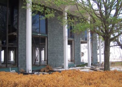 The exterior of a columned library building shows missing windows, damaged walls, and piles of debris at the base. Brown, dead shrubs line the walkway in front of the storm‑damaged structure.