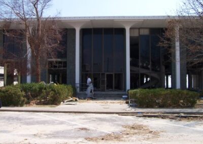 The front entrance of the Gulfport Library has shattered window panels and debris piled near damaged shrubs. A person in protective clothing stands at the doorway amid widespread exterior destruction.