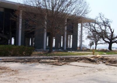 A wide view of the storm‑damaged Gulfport Library shows broken windows, debris covering the lawn and pavement, and several leafless trees framing the structure. The lower‑level interior is visible through missing walls and glass.