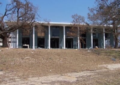 The Gulfport Library is viewed from a distance with its tall columned façade and bare, storm‑stripped trees. The surrounding ground is strewn with debris and dried vegetation from the hurricane surge.